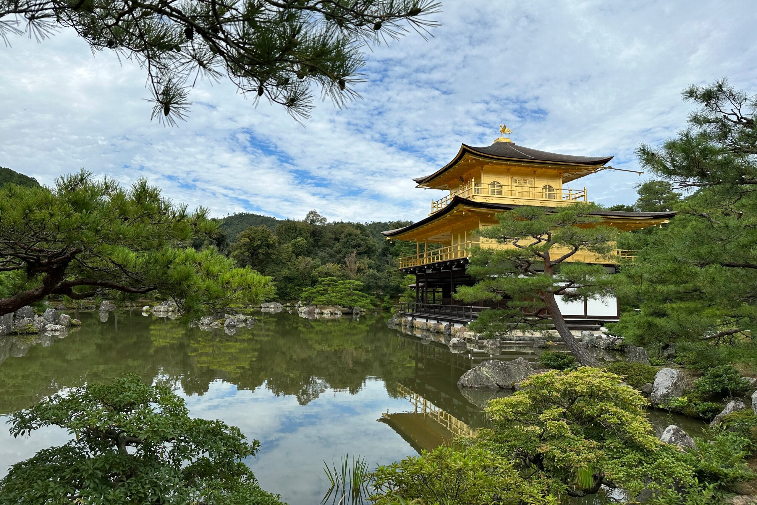 Der Goldene Pavillon (Kinkaku-ji) in Kyoto: dreistöckiger, goldverkleideter Tempel am Rand eines stillen Teichs. Deutliche Spiegelung im Wasser, Kiefern und Felsen im Vordergrund, blauer Himmel mit lockerer Bewölkung, keine Personen.