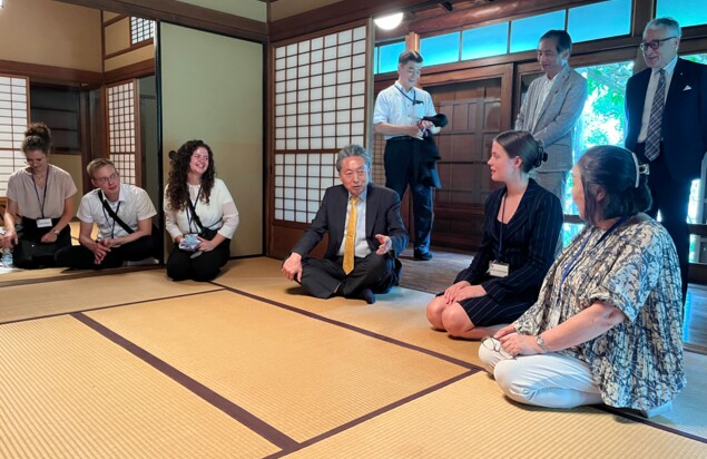 In a traditionally furnished Japanese house, several people are kneeling on tatami mats. Some are young and European, some older and Japanese. All are formally dressed. In the hallway behind the room, next to the open paper sliding doors, several more people can be seen standing.