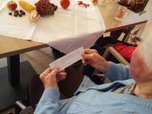 Eine Person im Rollstuhl hält einen Zettel mit einer vorgedruckten Fürbitte in den Händen. Auf dem Tisch liegt herbstliche Dekoration mit Kürbis, Kastanien, Mais und Äpfeln.