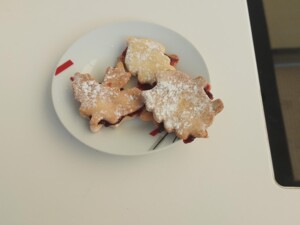Ein kleiner Teller mit drei herbstlich geformten Doppelkeksen, gefüllt mit Uhudlermarmelade und bestäubt mit Staubzucker, steht auf einem weißen Tisch.