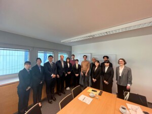 Zwölf Personen stehen in der Ecke eines Büroraums und posieren für ein Foto. Auf einer Wand sind Fenster, vor der Gruppe sind Schreibtische. Die Personen sind großteils formell gekleidet, was auf den offiziellen Charakter des Treffens deutet.