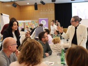 Mehrere Teilnehmende sitzen an einem Tisch, hören zu und diskutieren, während zwei Personen moderierend durch das Gespräch führen.