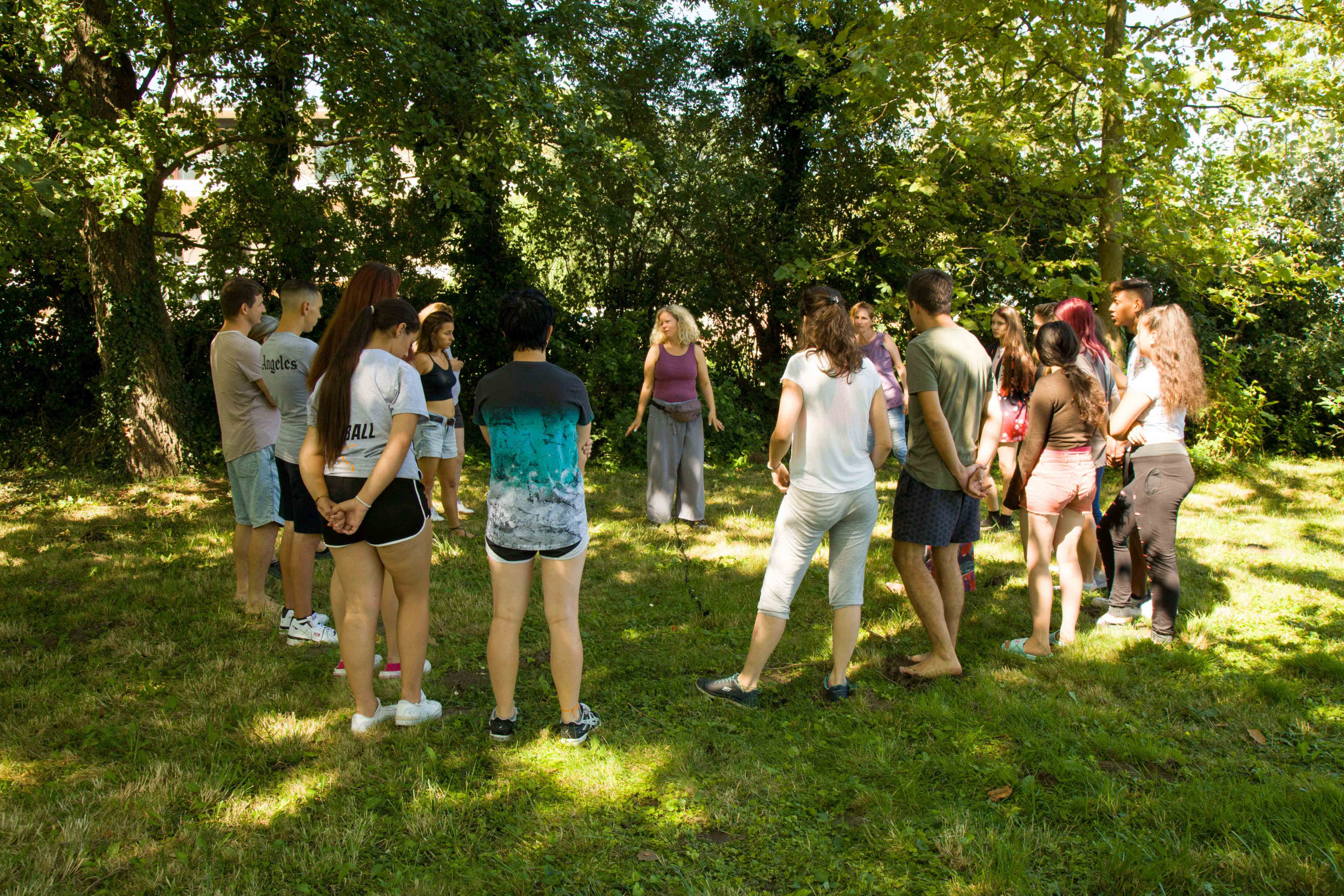 Jugendliche stehen auf einer grünen Wiese im Kreis und hören ihrer Ausbildnerin zu.