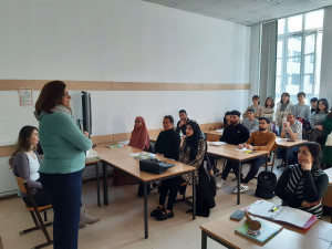 Schulunterrichtssituation mit SchülerInnen sitzend und im Hintergrund stehen und Lehrerin erklärend im Vordergrund stehend.