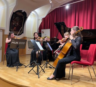 Vier Musiker:innen spielen gemeinsam Streichinstrumente in einem Ensemble. Sie sitzen auf Stühlen mit Notenständern vor sich. Im Hintergrund steht ein Flügel, und ein Schild mit der Aufschrift "die MUK" ist sichtbar.
