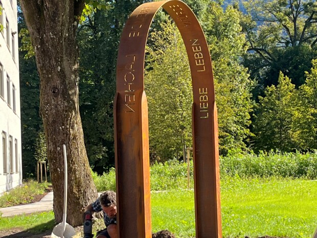 Ein vollständig montierter Bogen aus wetterfestem Stahl steht auf einem Weg zwischen Rasenflächen und Bäumen. Die Wörter "LIEBE", "LEBEN", "MUT", "WÜRDE", "FREUDE" und "LACHEN" sind entlang des oberen Bogens ausgestanzt. Im Hintergrund sind Berge und ein traditionelles Haus zu sehen. Foto: Birgit Schweiger.