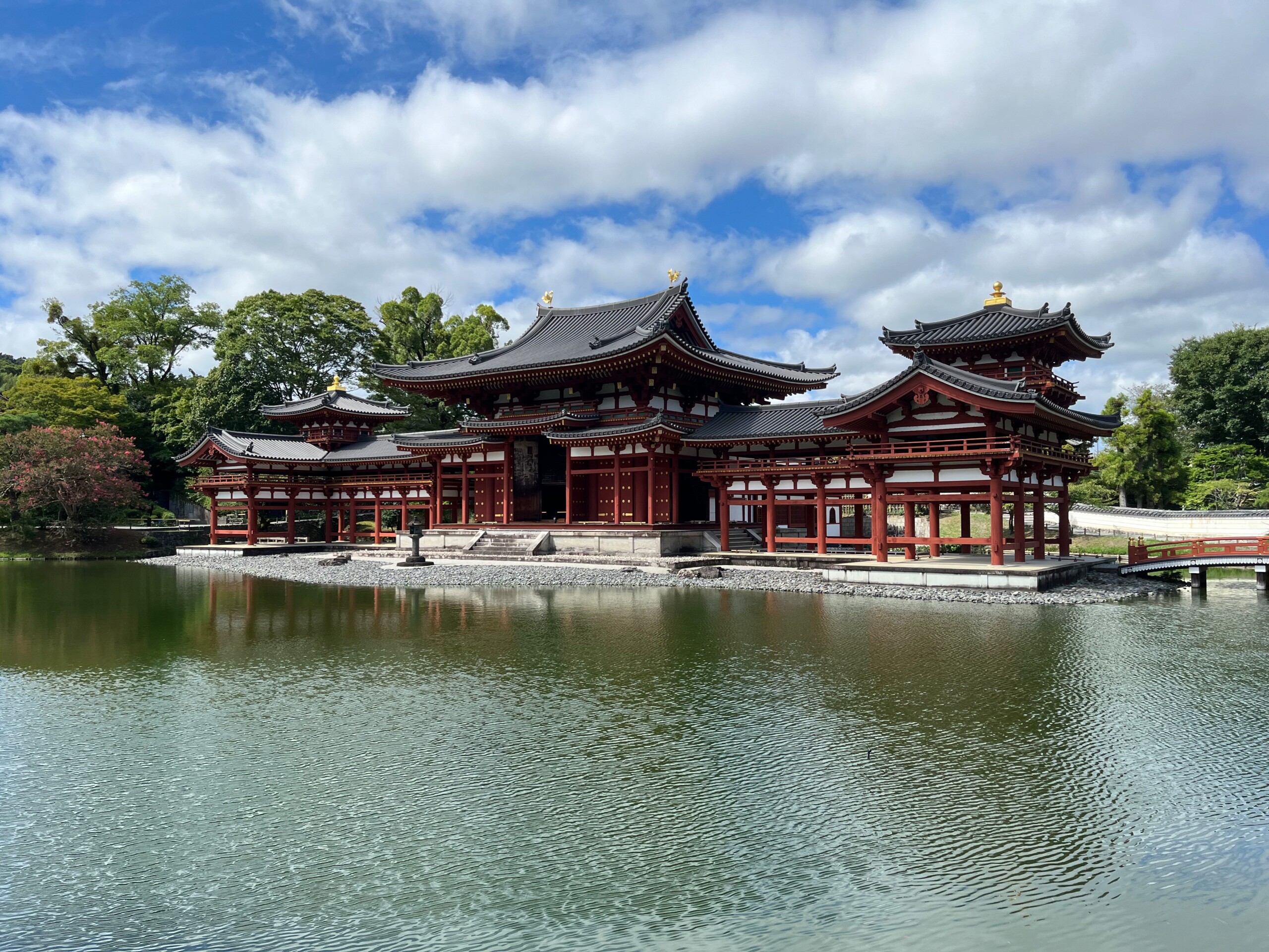 Schrägansicht auf den berühmten Tempel Byōdō-in in Kyoto: Weit ausladender Tempel mit geschwungenen, schwarzen Dächern und roten Holzsäulen. Der Tempel steht auf einer Insel in einem See und ist umringt von Bäumen.