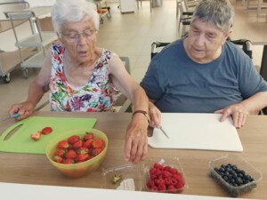 Zwei Bewohner:innen sitzen an einem Tisch und bereiten Obst für die Sommerbowle vor. Auf dem Tisch liegen Schneidebretter, Messer, Erdbeeren, Himbeeren und Heidelbeeren.