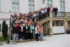 Große Gruppe von Teilnehmenden steht auf einer Treppe vor einem Gebäude und blickt in die Kamera für ein Erinnerungsfoto.