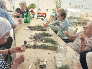 An einem großen Tisch sitzen mehrere ältere Personen, die Lavendelzweige zu kleinen Wichteln zusammenbinden. Auf dem Tisch liegen Scheren, Lavendelzweige und kleine Holzkugeln, die für die Köpfe der Wichtel verwendet werden. Die Stimmung ist konzentriert und entspannt.