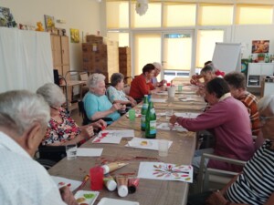 Mehrere ältere Menschen sitzen gemeinsam an einem langen Tisch und bemalen Papiere mit herbstlich gefärbten Baumkronen. Auf dem Tisch stehen Farben, Wasserbecher und Malutensilien.