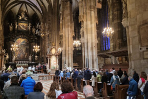 Zahlreiche Menschen nehmen an einem Gottesdienst im Stephansdom in Wien teil. Sie sitzen und stehen in den Kirchenbänken, während vorne Geistliche am Altar die Messe zelebrieren. Der gotische Innenraum ist reich mit Skulpturen, Altären und Gemälden verziert. Kronleuchter erhellen den Raum, und im Hintergrund ist ein Chor zu sehen.