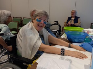 Eine Frau im Rollstuhl lächelt fröhlich in die Kamera, während sie eine Handpflege bekommt. Sie trägt blaue Augenpads. Andere Bewohner:innen sind ebenfalls im Raum und nehmen am Angebot teil.