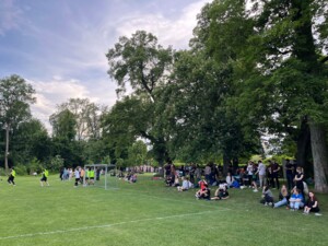 Zahlreiche Personen sitzen oder stehen am Spielfeldrand im Schatten von Bäumen und verfolgen ein Fußballspiel. Einige stehen auch hinter einem Tor auf der Wiese.