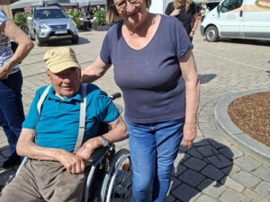 Eine Frau steht neben einem älteren Mann im Rollstuhl und legt ihm eine Hand auf die Schulter. Beide befinden sich auf einem sonnigen Marktplatz, umgeben von Gebäuden und weiteren Menschen.