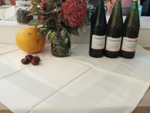 Ein herbstlich dekorierter Tisch mit drei Flaschen Traubensaft, einem gelben Kürbis, frischen Kastanien und einem Blumenstrauß mit Hortensien. Im Hintergrund steht eine Person in einem rosa Pullover.