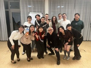 Mehrere Personen stehen und hocken in einem Innenraum mit Vorhängen im Hintergrund. Sie lehnen sich für ein gemeinsames Gruppenfoto zusammen, lächeln freundlich und blicken in die Kamera. Einige halten Getränke oder machen mit den Fingern ein Victory-Zeichen. In der Mitte steht eine ältere Person mit Brille, die ebenfalls lächelt. Die Atmosphäre ist warm, gesellig und familiär.