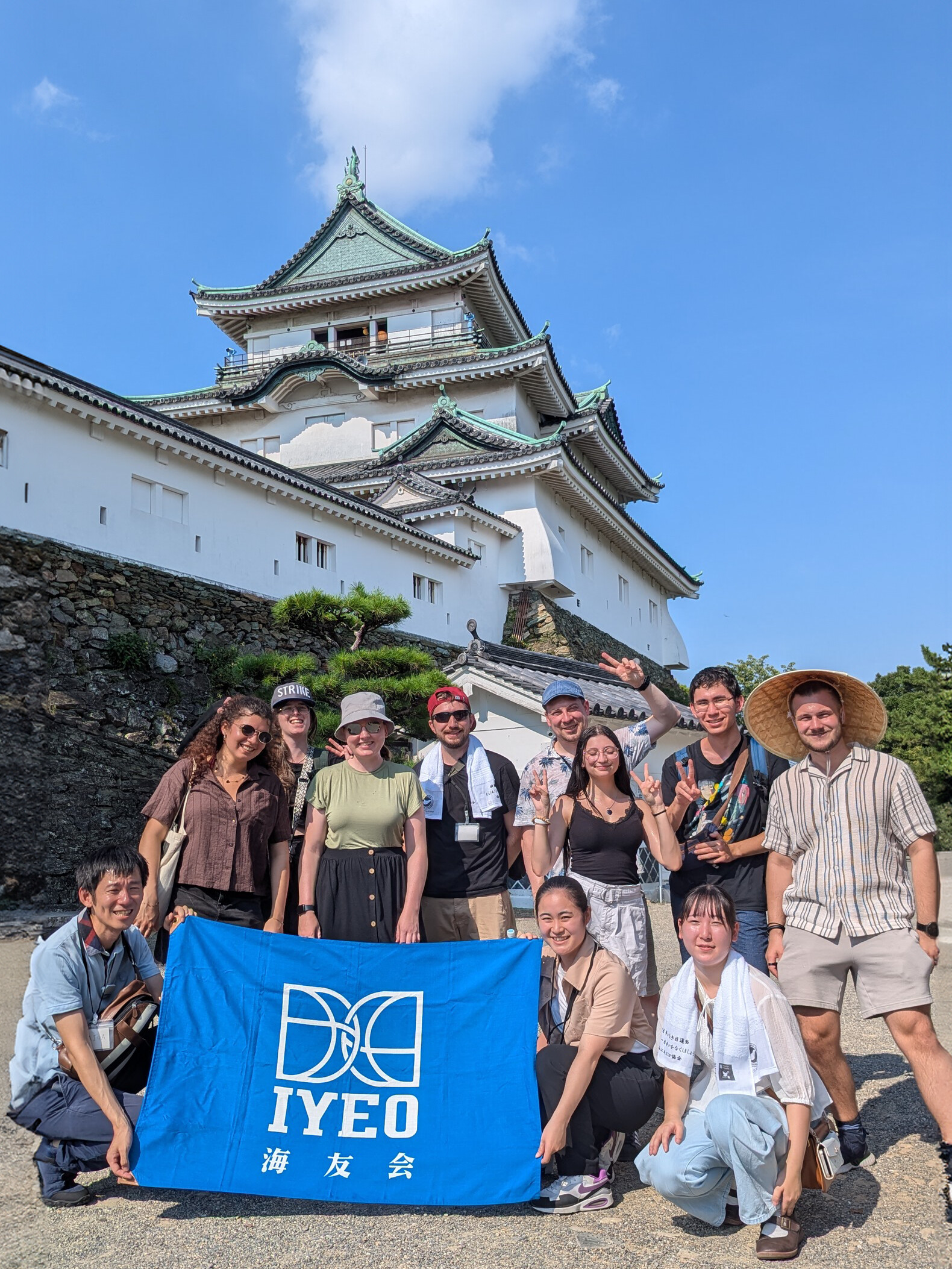 Eine gemischte Gruppe junger Menschen steht vor dem weißen Schloss Wakayama mit grünem Dach und hält ein blaues Banner mit der Aufschrift "IYEO". Der Himmel ist strahlend blau.