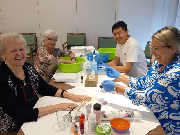 Vier Personen sitzen sich an einem Tisch gegenüber und lachen. Zwei von ihnen erhalten Handmassagen von Pflegekräften. Auf dem Tisch stehen Pflegeprodukte und Wasserschalen - eine entspannte Atmosphäre.