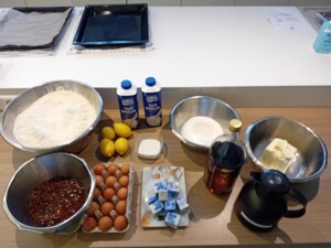 Auf einem Tisch stehen zahlreiche Backzutaten für Allerheiligen-Striezel bereit: Mehl, Eier, Rosinen, Zucker, Zitronen, frische Germwürfel, Butter, Milch, Rum, sowie eine schwarze Thermoskanne und zwei Backbleche im Hintergrund.