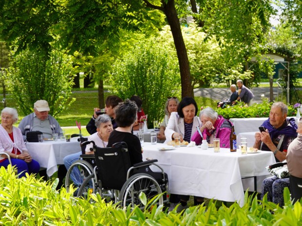 Eine Gruppe älterer Bewohner:innen, teils in Rollstühlen, sitzt mit Angehörigen an weiß gedeckten Tischen im schattigen Garten. Rundherum stehen grüne Sträucher und Bäume, auf den Tischen sind Getränke und kleine Speisen zu sehen.