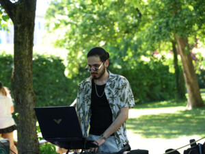 Ein DJ steht im Park unter Bäumen an einem Laptop und Musik-Equipment. Im Hintergrund sind grüne Hecken, Wiesen und weitere Besucher:innen des ÖJAB-Frühlingsfestes zu sehen.