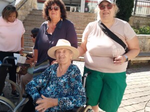 Eine Seniorin im Rollstuhl lächelt mit Sonnenhut in die Kamera; neben ihr stehen zwei Begleiter:innen. Im Hintergrund Marktstufen und Geschäfte. Aufnahme vom Marktausflug Anfang August in Güssing.