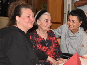 Drei Frauen sitzen dicht beisammen und lachen herzlich. Die ältere Dame in der Mitte trägt ein Kleid mit Rosenmuster und wird von zwei jüngeren Frauen begleitet.