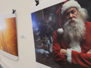 Ein Porträt eines digital generierten Santa Claus mit rotem Mantel, langem weißen Bart und verschränkten Armen. Der Hintergrund ist verschwommen, deutet jedoch eine Werkstatt-Umgebung an. Das Bild hängt in einer Ausstellung neben weiteren KI-Kunstwerken.