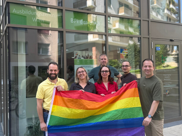 Mitarbeiter:innen der ÖJAB halten gemeinsam eine PRIDE-Flagge in den Händen, stehend vor dem Eingang zum BPI der ÖJAB in Wien.