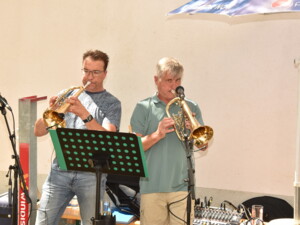 Zwei Männer stehen nebeneinander und spielen Trompeten. Vor ihnen steht ein Mikrofon und ein Notenständer. Beide konzentrieren sich auf ihre Instrumente.