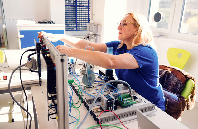 Eine Kursteilnehmerin in blauem Shirt sitzt an einem Arbeitstisch und justiert ein technisches Modell mit Kabeln, Schläuchen, Sensoren und Pneumatik-Bauteilen in einem hellen Schulungsraum. Foto: www.inframe.at