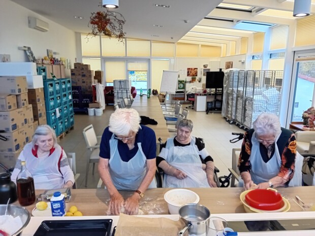 Vier Senior:innen sitzen und arbeiten an einem langen Tisch mit Backzutaten. Zwei von ihnen kneten Teig, während die anderen zusehen.