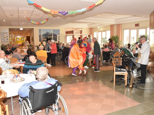 In der festlich geschmückten Eingangshalle des PFlegewohnhauses in Güssing sitzen zahlreiche Bewohner:innen an weiß gedeckten Tischen, viele nutzen Rollstühle oder Rollatoren. Bunte Girlanden hängen von der Decke. In der Mitte tanzen zwei als Clown verkleidete Ehrenamtliche mit einer Bewohnerin und einem Bewohner. Rechts spielen Musiker mit Trompete und Gesangsanlage. Die Atmosphäre wirkt fröhlich und lebendig, mehrere Personen beobachten das Geschehen oder unterhalten sich.