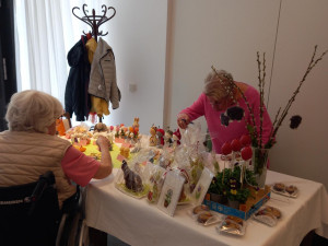 Zwei ältere Frauen, eine davon im Rollstuhl, stehen an einem dekorierten Stand mit Schokohasen, Karten, Osterblumen und Frühlingsartikeln.