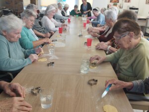 Eine große Gruppe älterer Menschen sitzt an einem langen Tisch und formt gemeinsam Kekse aus Teig. Verschiedene Ausstechformen und Teigstücke liegen auf dem Tisch.