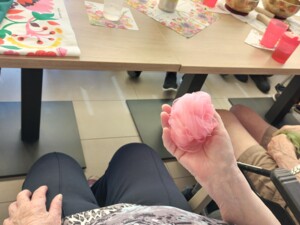 Nahaufnahme einer Hand, die ein rosa, leicht transparentes Tuch hält. Im Hintergrund sind der Tisch mit Blumendekoration, Bechern und Klangschalen sowie sitzende Teilnehmende zu sehen.