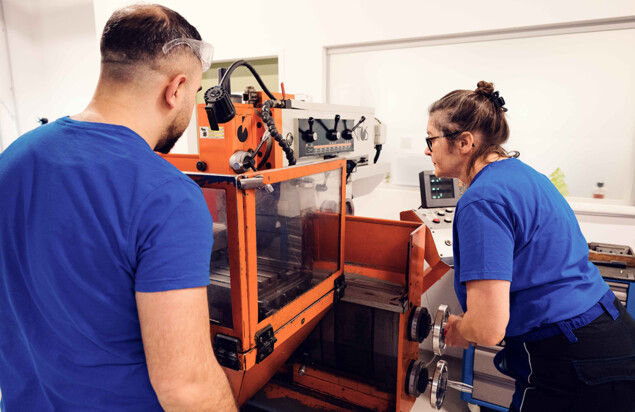 Zwei Kursteilnehmende in blauen Shirts arbeiten an einer großen orangefarbenen Industriemaschine. Eine Person steht links vor der Maschine, eine weitere bedient rechts ein Handrad und blickt auf die Steuerung. Foto: www.inframe.at 