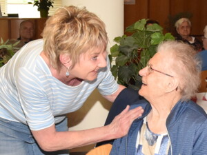 Eine Frau in einem gestreiften Shirt beugt sich zu einer älteren Dame im Rollstuhl. Beide blicken sich mit einem Lächeln an und halten Augenkontakt.
