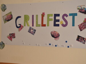 Plakat mit bunten Buchstaben "Grillfest", verziert mit Bildern von Grillgerichten, Obst und Blumen, hängt an einer Wand.