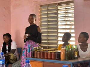 In einem Innenraum steht eine junge Frau vor einem Tisch und spricht zu einer kleinen Gruppe sitzender Personen. Auf dem Tisch sind zahlreiche Gläser mit eingemachten Produkten sowie Flaschen mit Getränken ordentlich aufgereiht. Die umstehenden Personen hören aufmerksam zu.