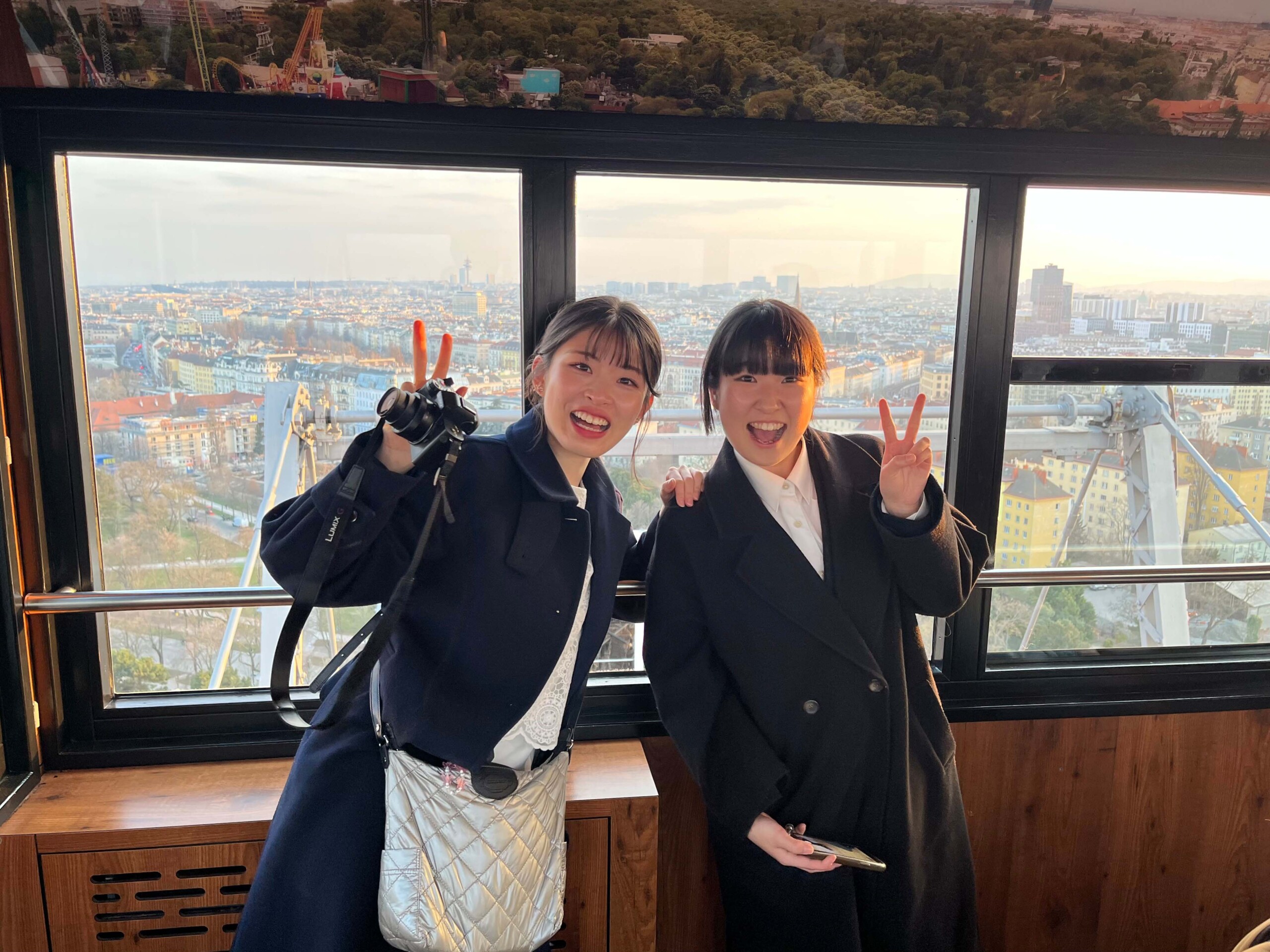 Two young Japanese women stand in a glass-enclosed observation gondola overlooking a sprawling city bathed in warm evening light. Both wear dark coats, smile at the camera, and each raises a hand in a victory sign. The woman on the left is also holding a camera, while in the background rooftops, buildings, and parts of the Ferris wheel or gondola structure are visible.