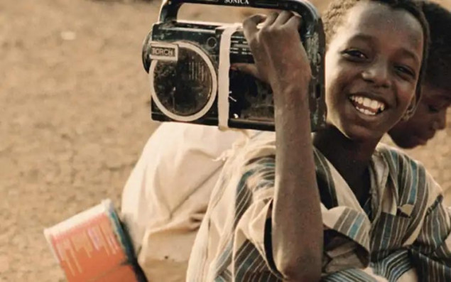 Symbolbild lächelndes afrikanisches Kind mit tragbarem Radio auf der Schulter im Vordergrund. Kind im Hintergrund sitzend.