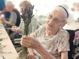 Eine ältere Bewohnerin hält einen Bündel frischer Lavendelzweige in der Hand und betrachtet ihn lächelnd. Im Hintergrund sind verschwommen weitere Bewohner:innen zu sehen, die ebenfalls Lavendelzweige bearbeiten.