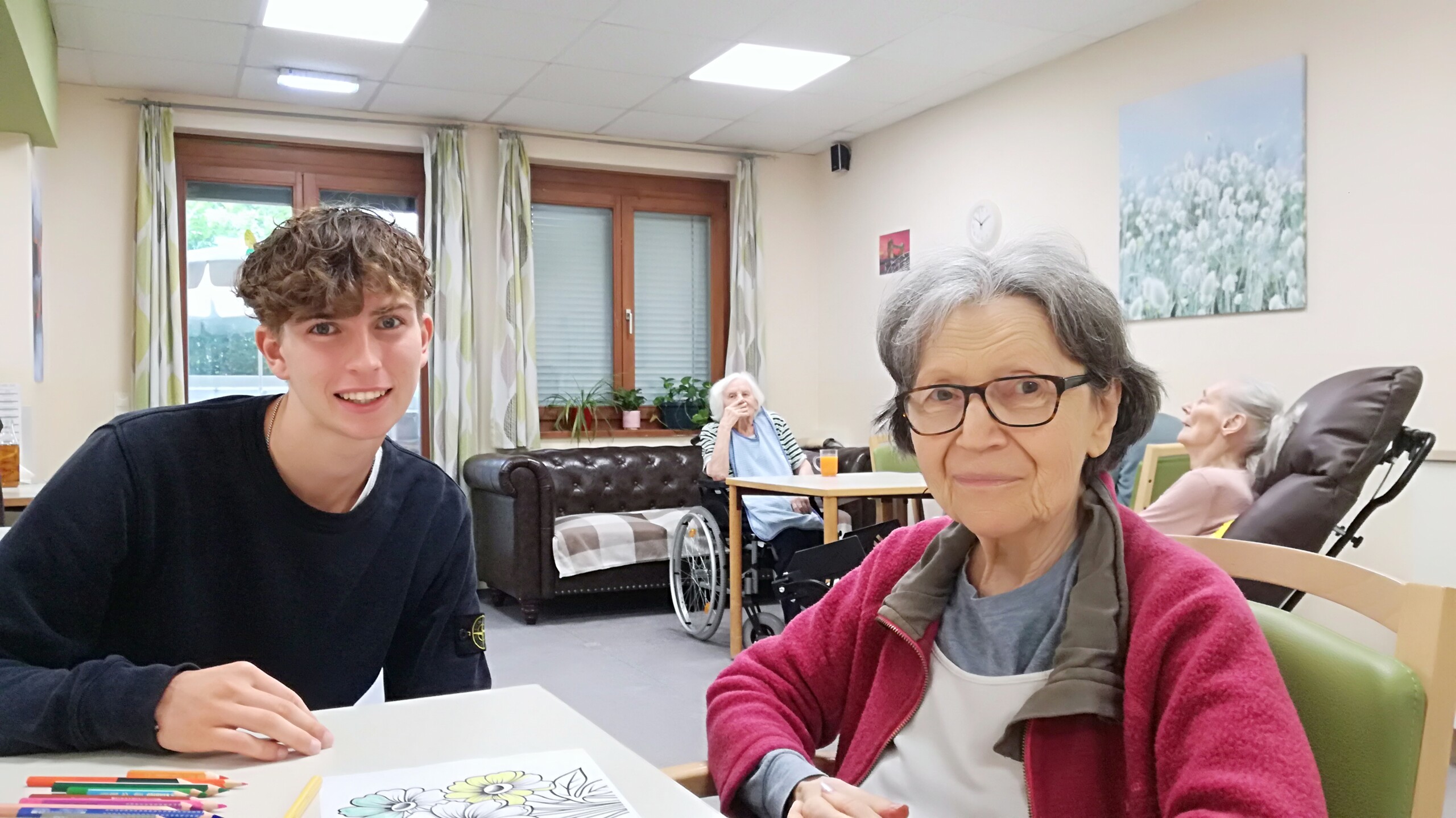 Ein junger Praktikant sitzt lächelnd neben einer älteren Bewohnerin in einem Pflegeheim. Auf dem Tisch vor ihnen liegt eine teilweise ausgemalte Zeichnung mit Buntstiften. Im Hintergrund sitzen weitere Senior:innen in gemütlicher Atmosphäre. Das Bild vermittelt einen Moment der Nähe und des Miteinanders im Alltag eines Sozialpraktikums.