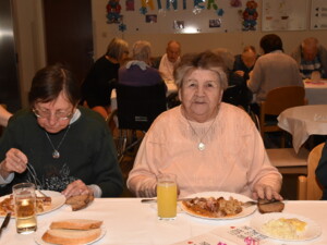Zwei ältere Frauen sitzen an einem Tisch. Eine isst konzentriert, die andere lächelt in die Kamera. Auf dem Tisch stehen Gläser mit Getränken und typische Speisen.