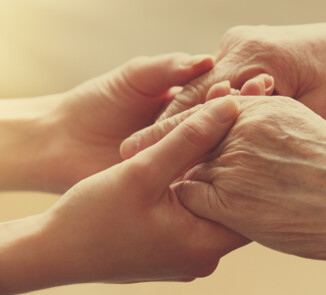 Eine jüngere Person hält sanft die Hände einer älteren Person. Die Szene ist in warmes Licht getaucht und vermittelt Nähe, Trost und gegenseitige Unterstützung.