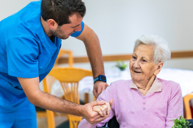 Symbolbild. Ein junger Pflegefachkraft in blauer Berufskleidung hält die Hand einer älteren Person im Rollstuhl und spricht ihr ermutigend zu. Die Seniorin trägt einen rosa Pullover und lächelt ihm zu. Im Hintergrund sind Tische und Stühle in einem hellen Aufenthaltsraum zu sehen.