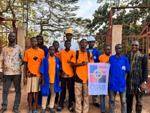 Eine Gruppe von Jugendlichen und erwachsenen Personen steht gemeinsam vor einem Eingangstor und blickt in die Kamera. Mehrere Personen tragen orangefarbene Shirts oder blaue Arbeitskittel, einige halten Schutzhelme oder tragen sie auf dem Kopf. In der Mitte wird ein Banner mit dem Logo der ÖJAB gehalten. Die Szene vermittelt Teamarbeit und gemeinschaftliches Engagement im Rahmen eines Ausbildungskontexts.