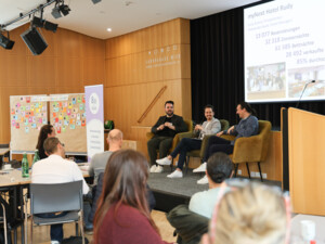 Drei Sprecher:innen sitzen auf einer Bühne und diskutieren vor Publikum; hinter ihnen wird eine Präsentation mit Zahlen und Bildern gezeigt.